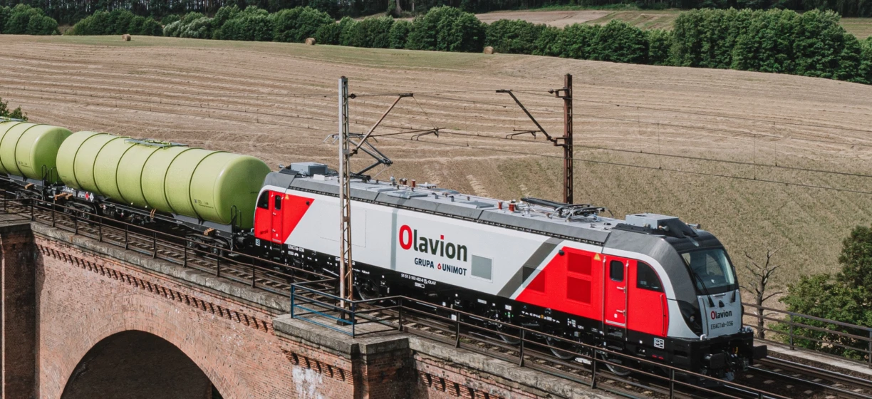 Lokomotywa elektryczna na torach kolejowych z innym taborami. Maszyna transportu publicznego firmy GRUPA & UNIMOT. Model Olavion S1 51 3 180 482-4 PL-CL.