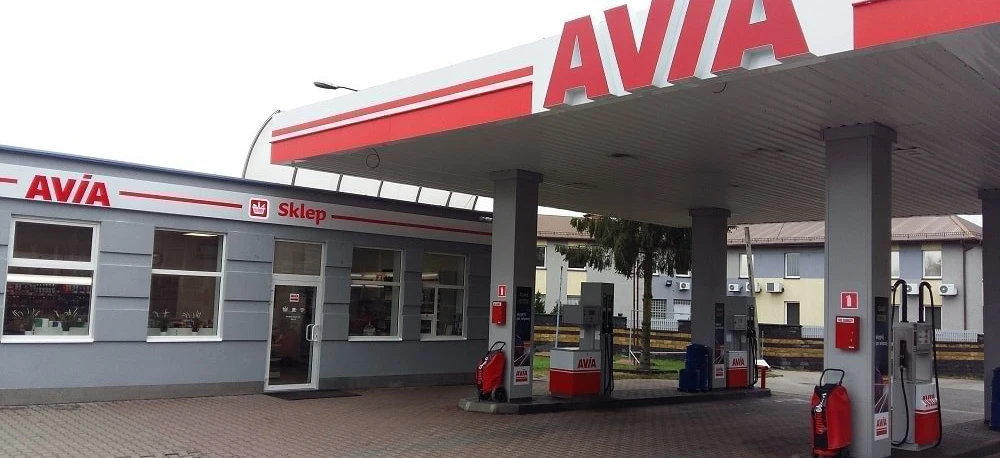 Zbliżenie dystrybutora paliwa na stacji benzynowej AVIA z lustrem samochodowym, zderzakiem i tablicą rejestracyjną w tle. - Stacja benzynowa AVIA.